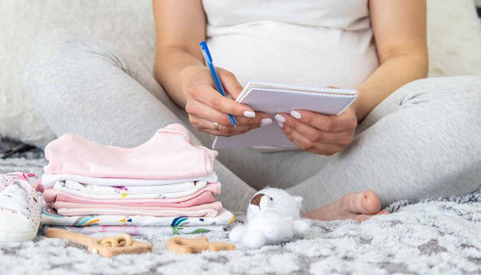 Pregnant woman writing a list of Baby Essentials while surrounded by baby clothes and toys.