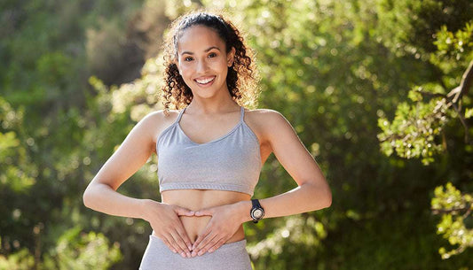 Smiling woman in athletic wear promoting gut health for weight loss by holding her stomach outdoors.
